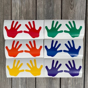 May include: A collection of colorful handprint decals in red, green, orange, blue, yellow, and purple. Each decal features a pair of open hands, perfect for decorating or crafting projects. The decals are arranged on a wooden surface.