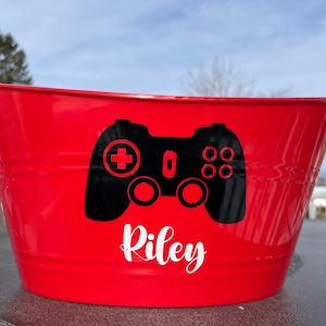 May include: Red metal bucket with a black video game controller and the name "Riley" in white lettering.