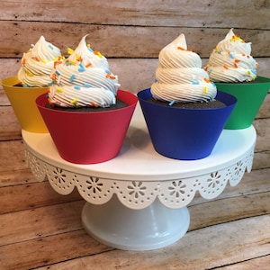 May include: Four cupcakes with white frosting and colorful sprinkles are displayed on a white cake stand with a lace trim. The cupcakes are in red, blue, and green cupcake liners.