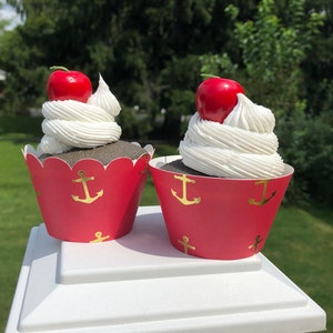 May include: Two red cupcake liners with gold anchor designs, each holding a chocolate cupcake topped with white frosting and a red cherry.