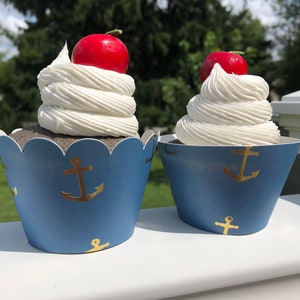 May include: Two blue and white cupcake liners with gold anchor designs. Each cupcake is topped with white frosting and a red cherry.