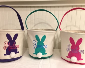 Puede incluir: Tres cestas de Pascua con siluetas de conejos y nombres personalizados. Las cestas son blancas con asas y ribetes morados, verde azulado y rosas. Los conejos tienen colas blancas y esponjosas. Los nombres son "Madison", "Jameson" y "Anderson".