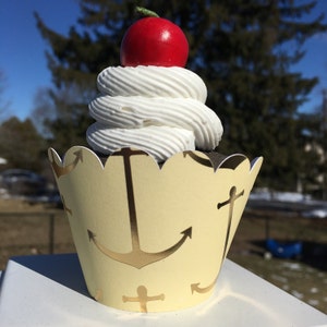 May include: A yellow cupcake liner with gold anchor designs and a white cupcake topped with white frosting and a red cherry.