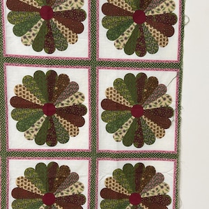May include: A fabric quilt top featuring six repeating floral designs. Each flower has a dark red center and petals in shades of green, brown, and cream. The flowers are set against a white background with a pink and green border.
