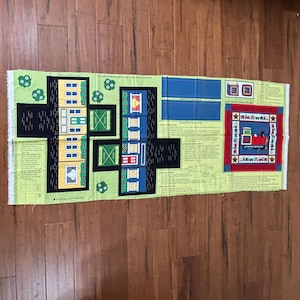 May include: A rectangular fabric panel featuring a colorful town scene with houses, trees, and a train. The houses are in various colors, including yellow, blue, and red. The panel includes instructions and is set against a green background.