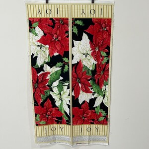 May include: Two fabric panels with a Christmas theme. The panels feature red and white poinsettias with green leaves against a black background. The word "JOY" is printed at the top of each panel. The bottom of each panel has text and a gold striped border.