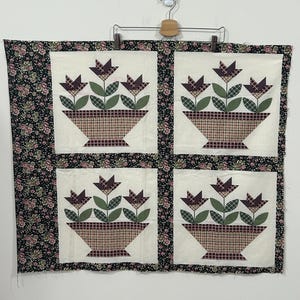 May include: A quilted wall hanging featuring four squares, each with a floral basket design. The baskets are plaid, and the flowers are purple. The border is black with a floral pattern. The piece is hung on a metal hanger.