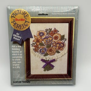 May include: A Bucilla "Picture Perfect" art stitchery kit, featuring a floral design titled "Lavender Nosegay." The kit includes a framed picture with a bouquet of flowers in shades of purple, orange, and brown. The kit's dimensions are 5 x 7 inches (12.7 x 17.8 cm).