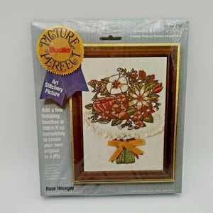 May include: A Bucilla "Picture Perfect" art stitchery kit featuring a floral design, framed in a brown wooden frame. The kit includes the materials to create a Rose Nosegay. The packaging includes text that reads "Art Stitchery Picture".