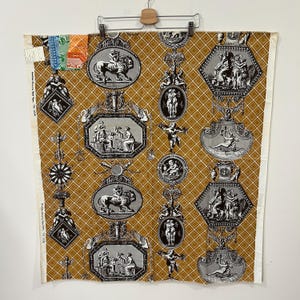 May include: A large piece of fabric with a repeating pattern of ornate medallions and figures in black and white against a gold background. The fabric is hanging from a wooden hanger. The medallions feature lions, figures, and classical scenes.