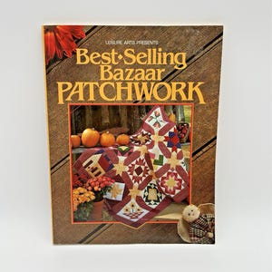 May include: A book cover titled "Best-Selling Bazaar Patchwork" by Leisure Arts. The cover features a patchwork quilt with star and floral designs, pumpkins, and fall foliage. The background is a wood-grain pattern.