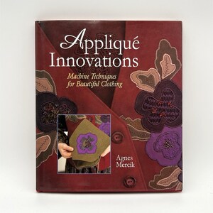 May include: A book titled "Appliqué Innovations" with the subtitle "Machine Techniques for Beautiful Clothing" by Agnes Mercik. The cover is burgundy with floral appliqué designs in shades of brown, purple, and black. A smaller image shows a person holding a piece of fabric with a floral design.