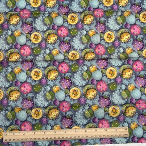 May include: Fabric with a repeating pattern of colorful, circular shapes in shades of blue, pink, yellow, green, and purple. The design includes a variety of sizes and textures, creating a visually interesting and dynamic pattern. A wooden ruler is at the bottom of the image.
