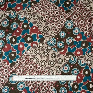 May include: A fabric with a repeating pattern of floral and geometric shapes in shades of brown, red, teal, and white. The design includes circles, dots, and stylized flower motifs. A ruler with text is at the bottom.