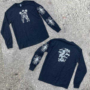 May include: Two black long-sleeved shirts with white graphic designs. One has a figure on the front and floral designs on the sleeves. The other has a bird design on the back and floral designs on the sleeves. The shirts are on a concrete surface.