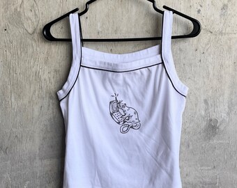May include: White vest top with black trim and straps, hanging on a black hanger. The top features a graphic of a cat holding a phone. The vest top is made of a soft, lightweight material.