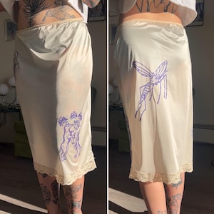 May include: Cream-colored slip skirt with a purple insect and animal graphic print. The skirt has a lace trim at the hem and a satin finish. The skirt is knee-length and has a high waist.