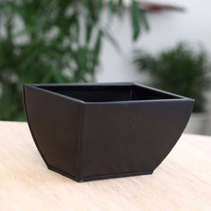 Puede incluir: Un macetero de metal negro con forma cuadrada y bordes redondeados. El macetero está hecho de un material duradero y es perfecto para uso en interiores o exteriores.