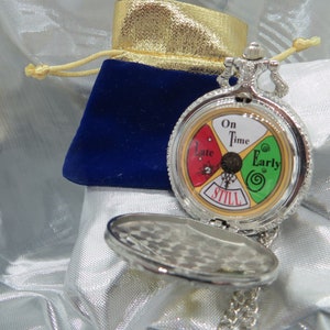 May include: Silver-toned pocket watch with a colorful face indicating time status: "On Time," "Early," "Late," and "Still." The watch is open, revealing the detailed face. A blue velvet pouch with a gold drawstring is in the background.