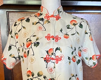 Pode incluir: Uma camisa de manga curta de cor creme com colarinho mandarim e estampa de fruta de caqui. A camisa tem botões vermelhos atados e uma estampa de caracteres chineses dourados.