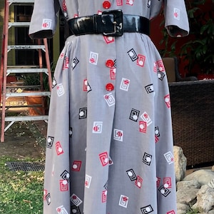 Könnte beinhalten: Graues Knopfkleid mit schwarzem Gürtel. Das Kleid hat ein sich wiederholendes Muster aus roten, schwarzen und weißen geometrischen Formen. Rote Knöpfe zieren die Vorderseite. Das Kleid wird auf einer Schaufensterpuppe präsentiert.