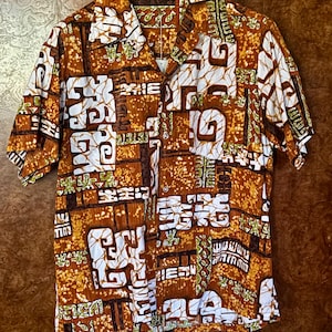 Peut inclure: Chemise à manches courtes avec un motif géométrique marron, blanc et vert. La chemise a un col et est suspendue à un cintre noir. Le tissu a un aspect texturé.