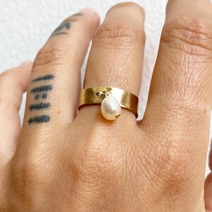 May include: A gold ring with a small pearl dangling from it. The ring is worn on a finger with a tattoo.