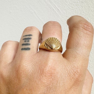 May include: A gold ring featuring a seashell design. The ring is worn on a finger and has a detailed, textured surface. The seashell design is the focal point, with radiating lines emanating from a central point.