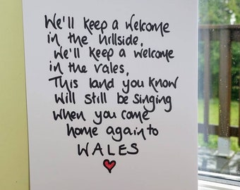 May include: A white paper print with handwritten black text. The text reads "We'll keep a welcome in the hillside...home again to WALES" with a red heart at the bottom. The print is displayed near a window.