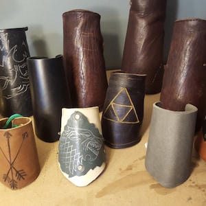 May include: A collection of 11 leather arm guards with various designs, including a wolf, arrows, a triangle, and studs. The arm guards are made of brown, black, and gray leather.