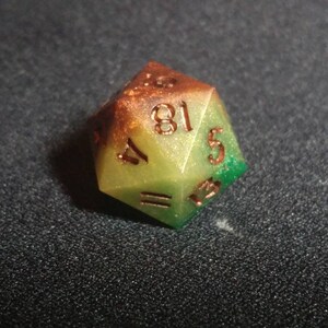 Puede incluir: Un dado de veinte caras con un degradado de color que va desde un marr&oacute;n &aacute;mbar intenso en la parte superior hasta un verde lima vibrante en la base. El dado tiene un acabado ligeramente brillante y n&uacute;meros grabados en un tono cobre met&aacute;lico.