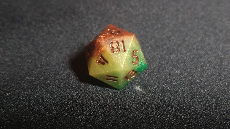 Puede incluir: Un dado de veinte caras con un degradado de color que va desde un marr&oacute;n &aacute;mbar intenso en la parte superior hasta un verde lima vibrante en la base. El dado tiene un acabado ligeramente brillante y n&uacute;meros grabados en un tono cobre met&aacute;lico.