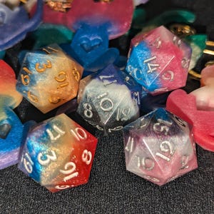Può includere: Un set di sei dadi iridescenti con numeri bianchi. I dadi sono realizzati in resina e hanno un aspetto maculato, simile a una galassia. I dadi sono di vari colori, tra cui blu, rosa, viola e arancione.