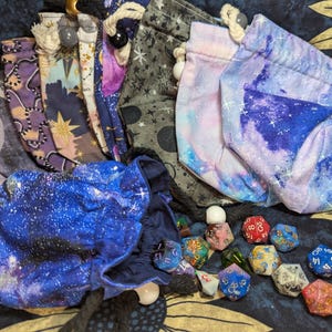May include: An assortment of dice bags and dice. The bags feature celestial patterns in shades of blue, purple, and gray. Dice are scattered around the bags, with various colors and designs. The bags have drawstring closures.