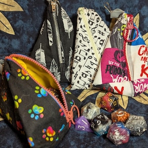 May include: Four triangular zippered pouches with different patterns: black with feathers, white with musical notes, white with a pink and black print, and black with colorful paw prints.  The pouches are arranged on a blue and gold patterned fabric with six colorful dice.