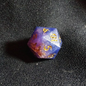 Puede incluir: Un dado de veinte caras con cuerpo de resina transl&uacute;cida en tonos morado y azul con escamas doradas incrustadas. Los n&uacute;meros est&aacute;n grabados en un acabado dorado, mostrando claramente los d&iacute;gitos 4, 18, 5 y 15 en sus caras visibles.