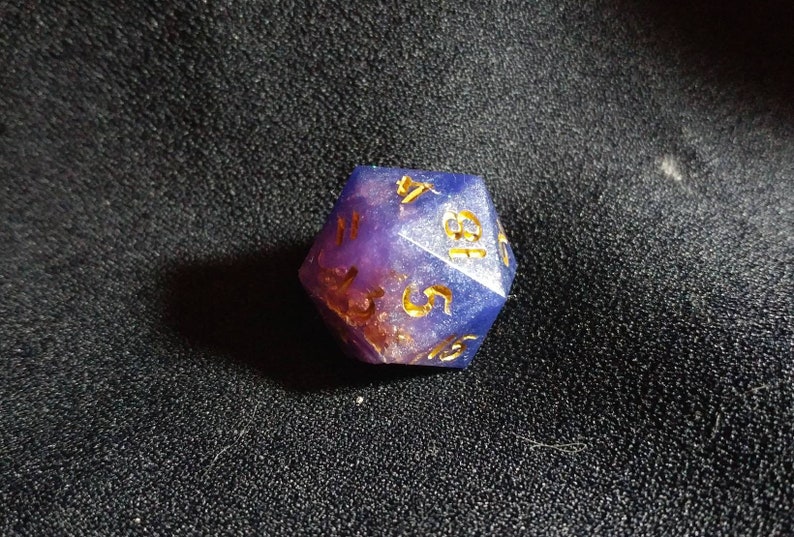 Puede incluir: Un dado de veinte caras con cuerpo de resina transl&uacute;cida en tonos morado y azul con escamas doradas incrustadas. Los n&uacute;meros est&aacute;n grabados en un acabado dorado, mostrando claramente los d&iacute;gitos 4, 18, 5 y 15 en sus caras visibles.