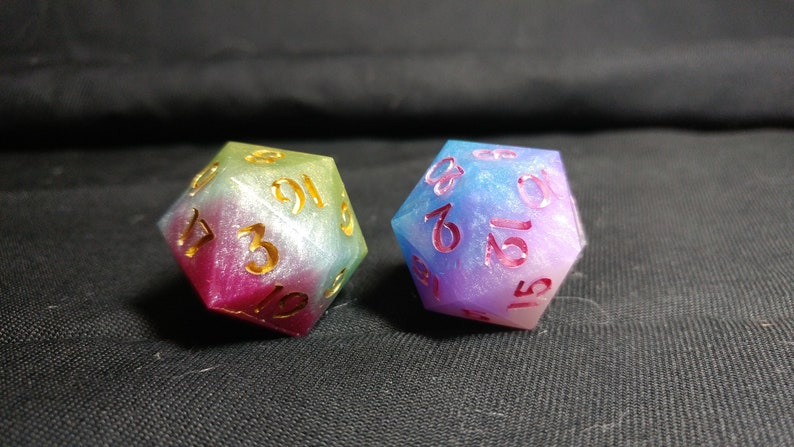 May include: Two iridescent dice with gold numbers. The dice are a gradient of pink, blue, and green.