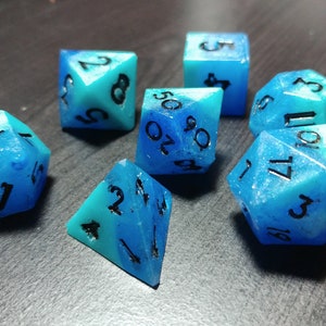 Puede incluir: Un conjunto de siete dados poli&eacute;dricos en tonos azules y turquesas. Cada dado presenta n&uacute;meros negros y un aspecto transl&uacute;cido veteado. Estos dados est&aacute;n dise&ntilde;ados para juegos de rol de mesa.