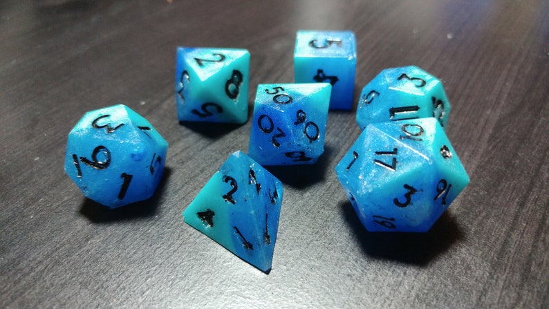 Puede incluir: Un conjunto de siete dados poli&eacute;dricos en tonos azules y turquesas. Cada dado presenta n&uacute;meros negros y un aspecto transl&uacute;cido veteado. Estos dados est&aacute;n dise&ntilde;ados para juegos de rol de mesa.