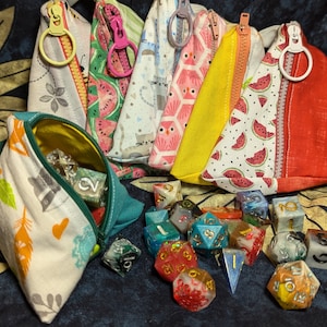May include: A set of six colorful fabric pouches with zipper closures and a variety of patterns, including watermelon, cats, and geometric designs. The pouches are arranged in a row on a dark floral fabric background.  A collection of colorful dice are scattered in front of the pouches.