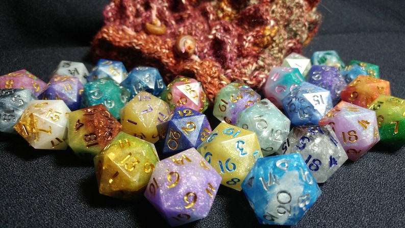 Puede incluir: Una colecci&oacute;n de dados multicolores con n&uacute;meros dorados y azules. Los dados son de varios colores, incluyendo azul, verde, morado y amarillo. Un tejido de punto est&aacute; en el fondo.