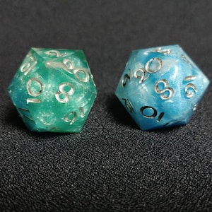 Puede incluir: Dos dados de veinte caras, uno en verde esmeralda transl&uacute;cido y el otro en azul cielo transl&uacute;cido, ambos con un brillo nacarado en el material de resina y n&uacute;meros grabados de color plateado en cada una de las caras triangulares.