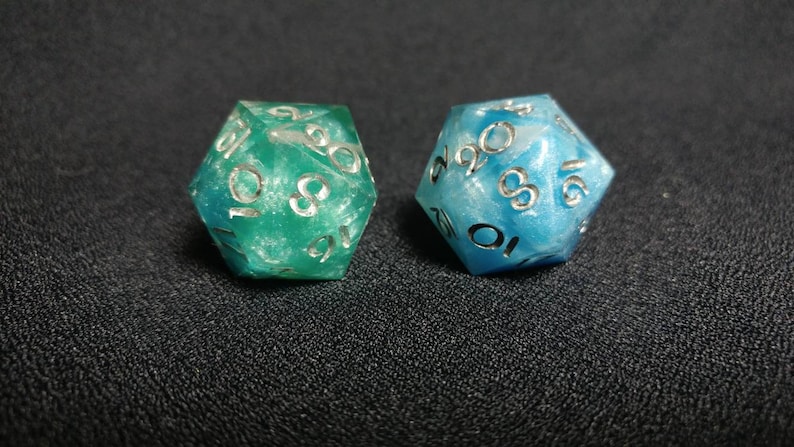 Puede incluir: Dos dados de veinte caras, uno en verde esmeralda transl&uacute;cido y el otro en azul cielo transl&uacute;cido, ambos con un brillo nacarado en el material de resina y n&uacute;meros grabados de color plateado en cada una de las caras triangulares.