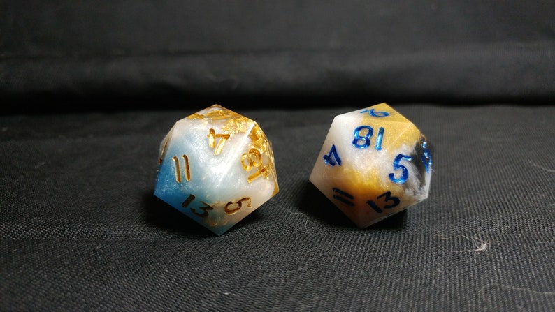 May include: Two white, blue, and gold dice with black numbers. The dice are made of resin and have a cloudy, marbled appearance.