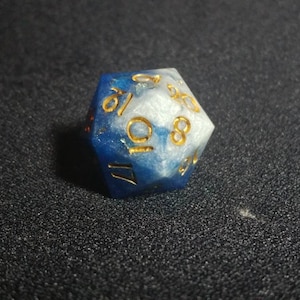 Puede incluir: Un dado de veinte caras que presenta una mezcla arremolinada de resina azul profundo y blanco nacarado. Los n&uacute;meros est&aacute;n grabados en la superficie del dado y rellenos con un pigmento dorado met&aacute;lico, ofreciendo un contraste claro con los colores.