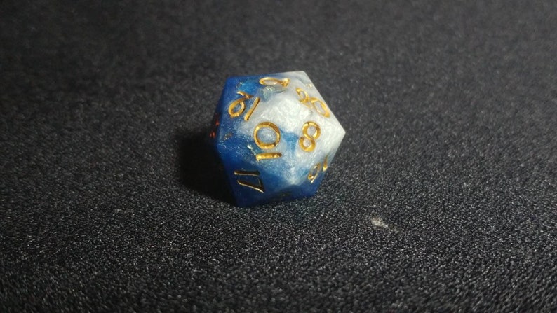 Puede incluir: Un dado de veinte caras que presenta una mezcla arremolinada de resina azul profundo y blanco nacarado. Los n&uacute;meros est&aacute;n grabados en la superficie del dado y rellenos con un pigmento dorado met&aacute;lico, ofreciendo un contraste claro con los colores.