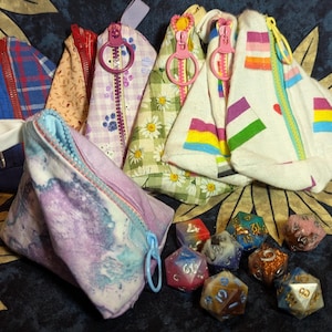 May include: A collection of nine colorful fabric pouches with zipper closures. The pouches are made of different fabrics and have various patterns, including plaid, floral, and geometric designs. The pouches are arranged on a dark blue background with a few dice scattered around them.