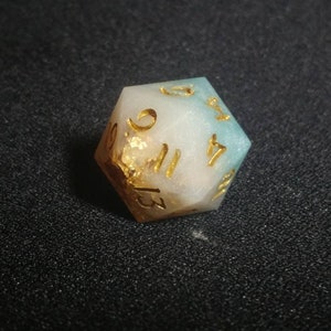 Puede incluir: Un dado de veinte caras con un degradado de resina blanca opaca y azul verdoso suave. El dado incluye escamas de color dorado incrustadas y n&uacute;meros grabados rellenos de tinta dorada, mostrando claramente los n&uacute;meros 9, 11, 13 y 17.