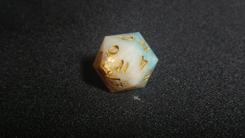Puede incluir: Un dado de veinte caras con un degradado de resina blanca opaca y azul verdoso suave. El dado incluye escamas de color dorado incrustadas y n&uacute;meros grabados rellenos de tinta dorada, mostrando claramente los n&uacute;meros 9, 11, 13 y 17.